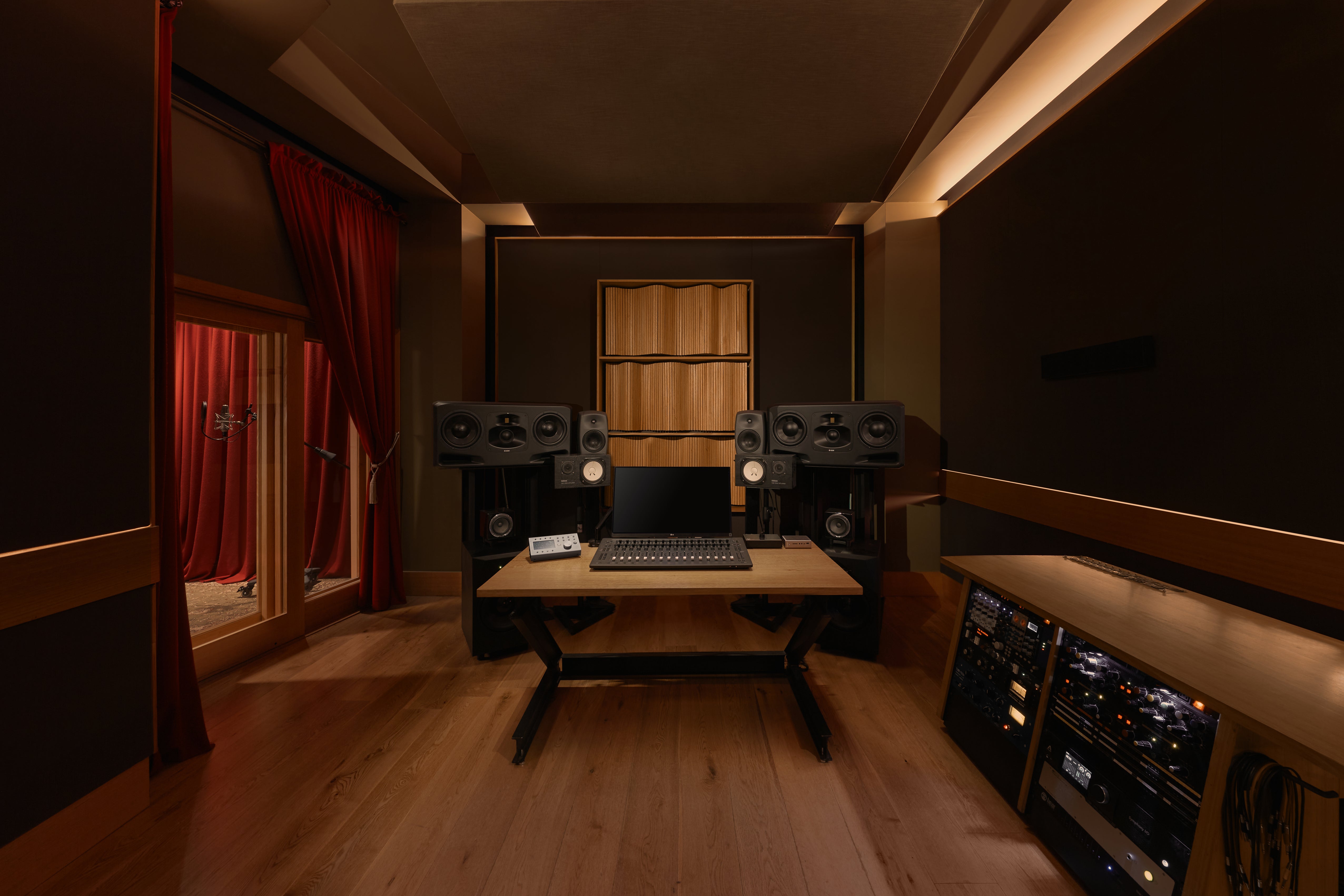 Professional recording studio setup featuring high-end audio monitors, sound-dampening wooden acoustic panels, a central mixing desk, and modern audio equipment. Dimly lit ambiance with red curtains and wooden flooring for optimal sound insulation